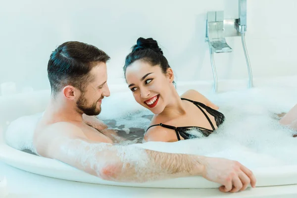 Hombre sin camisa guapo acostado en la bañera y mirando alegre morena mujer en sujetador negro - foto de stock