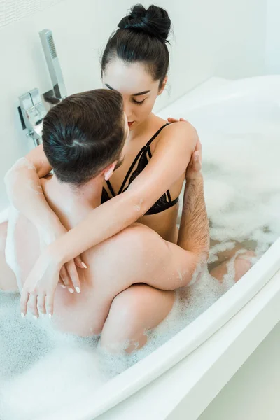 Back view of man hugging sexy brunette woman in black bra while sitting in bathtub with bath foam — Stock Photo