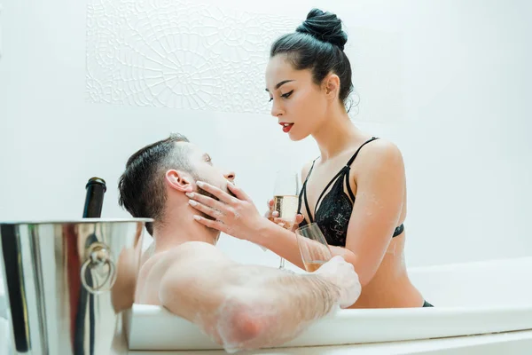 Sexy young woman holding champagne glass and touching face of handsome man in bathtub — Stock Photo