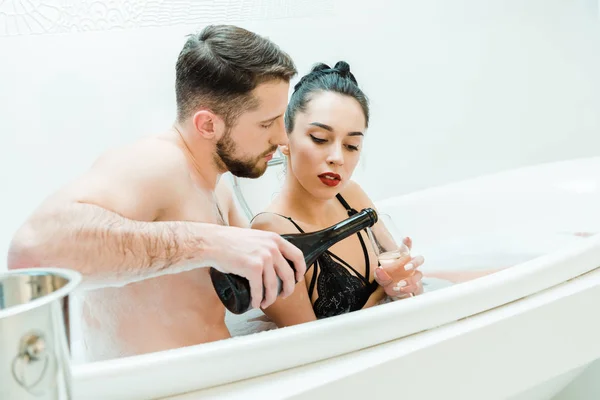 Homem bonito segurando garrafa perto de copo de champanhe de mulher na banheira — Fotografia de Stock