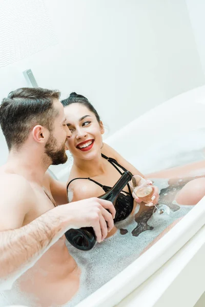 Весёлый бородатый мужчина держит бутылку возле бокала шампанского счастливой женщины в ванной — стоковое фото