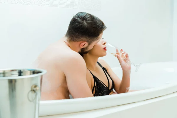 Селективное внимание мужчины целующего щеку молодой женщины, пьющей из бокала шампанского в ванной — стоковое фото