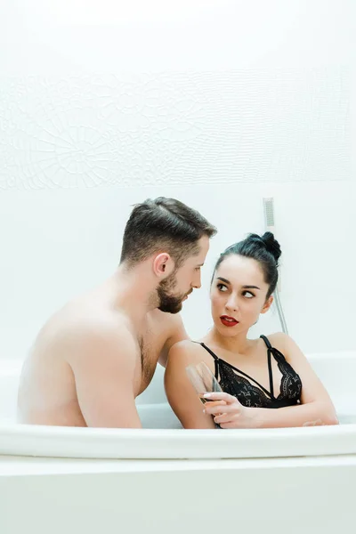 Hombre sin camisa guapo mirando mujer morena sosteniendo copa de champán en la bañera - foto de stock