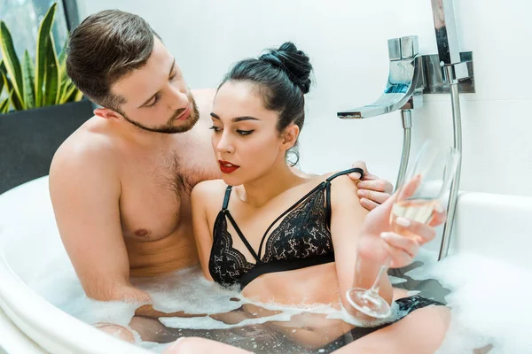 Barbuto senza maglietta uomo spogliarsi bruna donna in possesso di bicchiere di champagne nella vasca da bagno — Foto stock