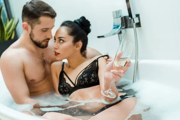Selektiver Fokus der brünetten Frau, die ein Champagnerglas hält und den Mann in der Badewanne ansieht — Stockfoto