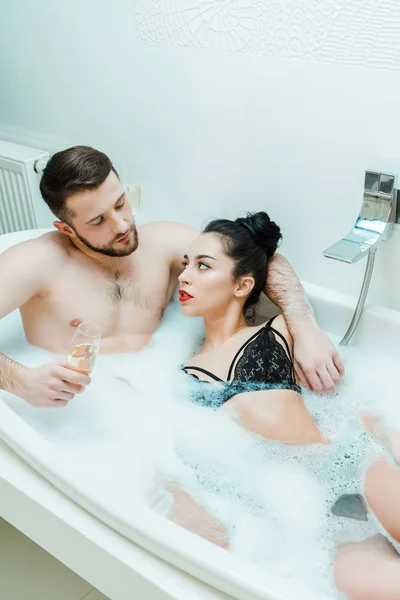 Shirtless man hugging brunette woman and holding champagne glass in bathtub — Stock Photo