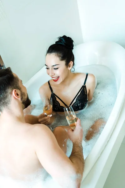 Vista aérea de la chica feliz mirando al hombre mientras sostiene la copa de champán en la bañera - foto de stock