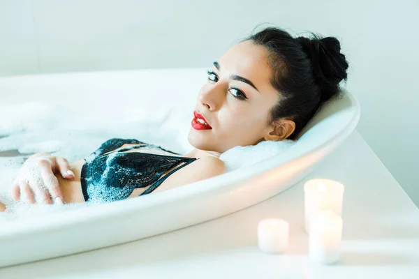 Attrayant brunette jeune femme regardant la caméra tout en étant couché dans la baignoire avec mousse de bain — Photo de stock