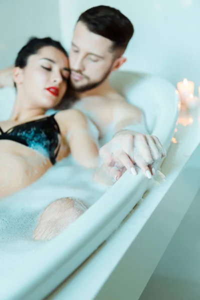 Messa a fuoco selettiva di uomo barbuto e attraente donna bruna che si tiene per mano mentre si trova nella vasca da bagno con schiuma da bagno — Foto stock