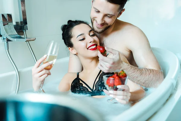 Messa a fuoco selettiva di felice giovane donna che tiene bicchiere di champagne e guardando gustosa fragola vicino fidanzato allegro nella vasca da bagno — Foto stock