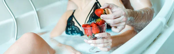 Panoramic shot of man holding tasty strawberry near woman in black bra lying in bathtub — Stock Photo