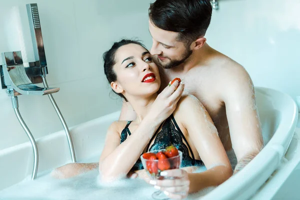 Bel homme regardant la femme attrayante tenant savoureuse fraise dans la baignoire — Photo de stock