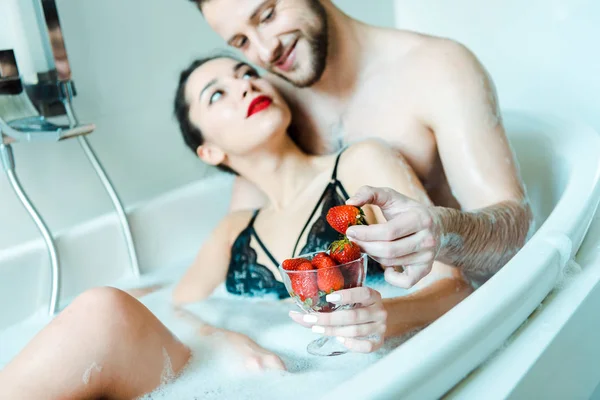 Selective focus of man holding tasty strawberry near woman in black bra lying in bathtub — Stock Photo