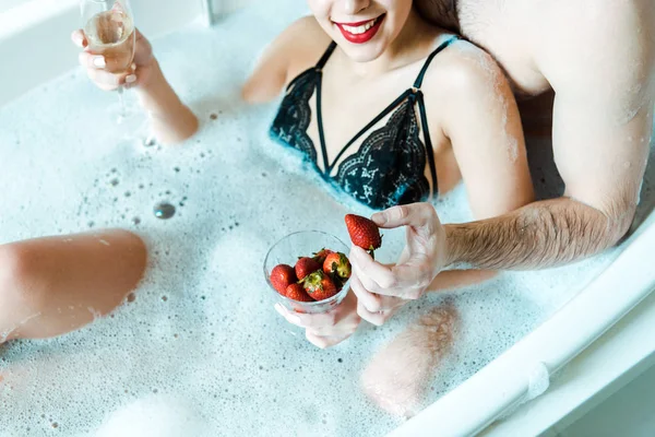 Abgeschnittene Ansicht eines Mannes, der leckere Erdbeeren in der Nähe eines glücklichen Mädchens mit Champagnerglas in der Badewanne hält — Stockfoto