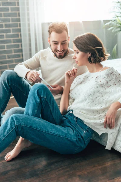 Alegre hombre sosteniendo teléfono inteligente cerca atractiva morena mujer en casa - foto de stock