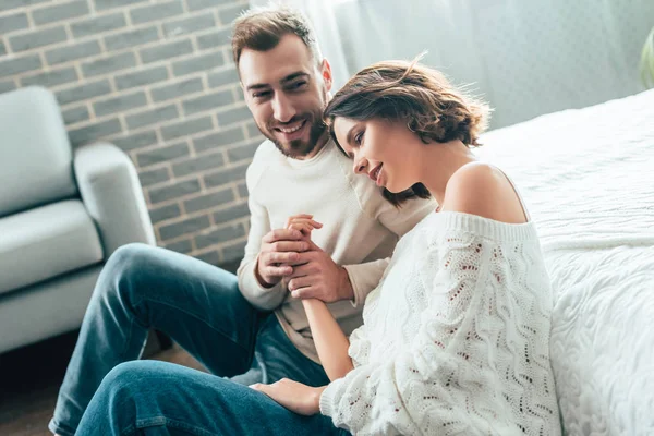 Hombre feliz cogido de la mano con novia atractiva mientras está sentado en el suelo en casa - foto de stock