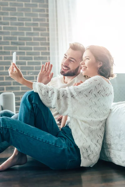 Mulher feliz acenando a mão enquanto toma selfie com o homem em casa — Fotografia de Stock