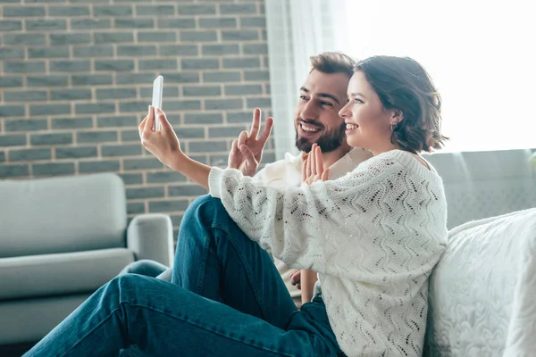 Happy woman waving hand while taking selfie with man showing peace sign at home — Stock Photo