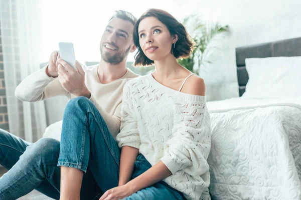 Glücklicher Mann mit Smartphone neben verträumter brünetter Frau zu Hause — Stockfoto