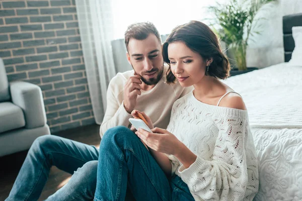 Mujer atractiva mirando el teléfono inteligente cerca de hombre guapo en casa - foto de stock