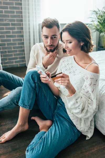 Femme gaie avec pieds nus assis sur le sol près de l'homme et tenant smartphone et carte de crédit — Photo de stock