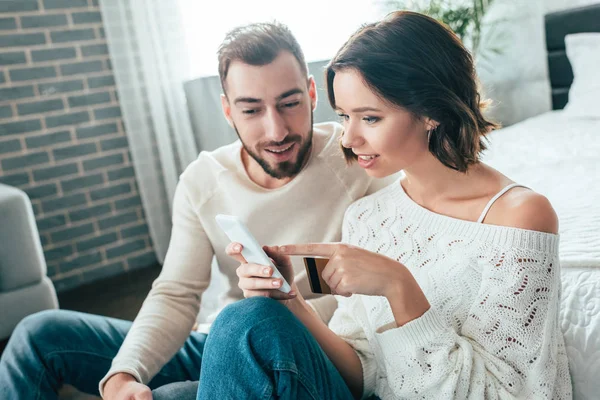 Atractiva mujer señalando con el dedo en el teléfono inteligente y la celebración de la tarjeta de crédito cerca del hombre feliz - foto de stock