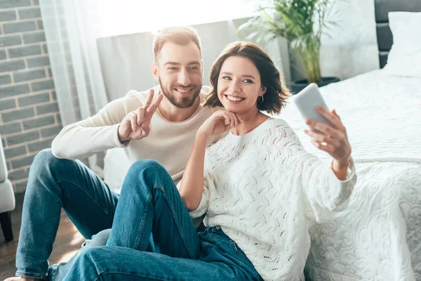 Selektiver Fokus der attraktiven Frau, die ein Selfie mit einem hübschen Mann macht, der ein Friedenszeichen zeigt — Stockfoto