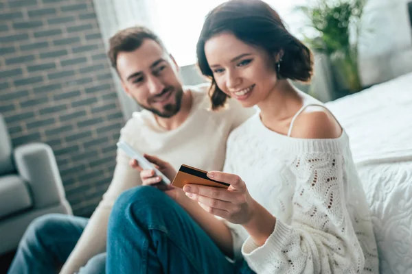 Foyer sélectif de femme heureuse tenant smartphone et regardant la carte de crédit près de l'homme joyeux — Photo de stock