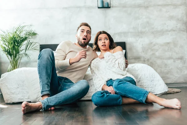 Sorprendido hombre y mujer sentado en el suelo y viendo la película en casa - foto de stock