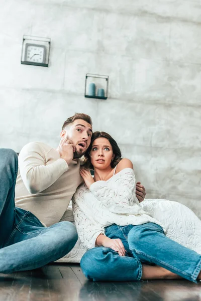 Vista de ángulo bajo de la pareja sorprendida sentada en el suelo y viendo la película en casa - foto de stock