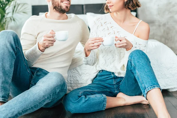 Cropped view of man and woman sitting on floor and holding cups — Stock Photo