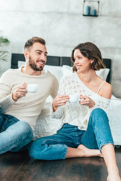 Happy man and woman sitting on floor and holding cups with drinks — Stock Photo