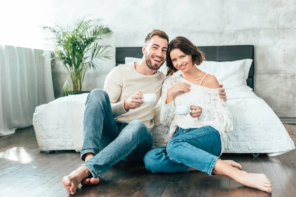 Handsome man hugging beautiful woman and sitting on floor with cup — Stock Photo