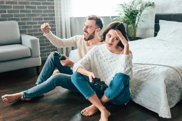 Happy man celebrating triumph near upset girl sitting on floor with joystick — Stock Photo