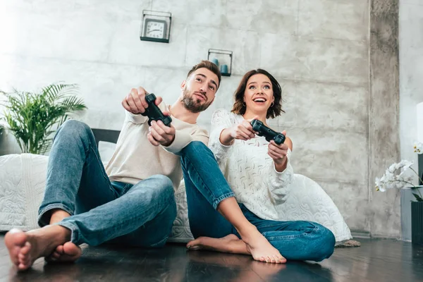 Low angle view of happy man and cheerful woman holding joysticks at home — Stock Photo