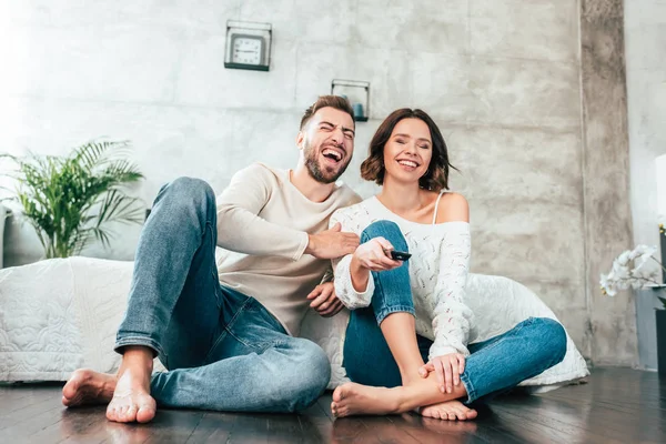 Baixo ângulo de visão do homem feliz sentado no chão perto de mulher alegre com controlador remoto — Fotografia de Stock