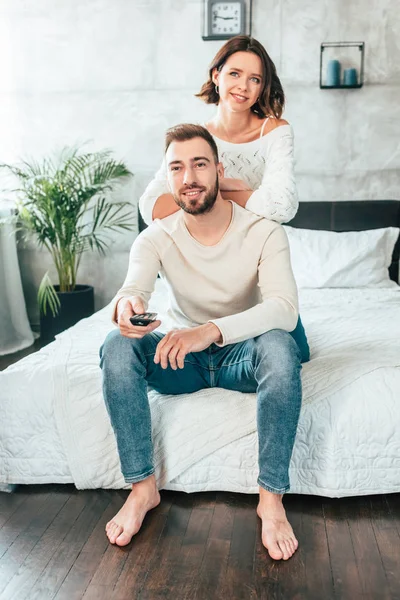 Mujer feliz cerca de hombre guapo sentado en la cama y sosteniendo el mando a distancia - foto de stock