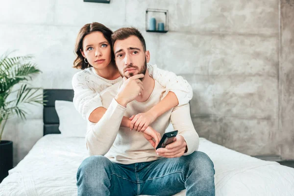Attraktive Frau umarmt aufgebrachten Mann, der auf Bett sitzt und Fernbedienung in der Hand hält — Stockfoto