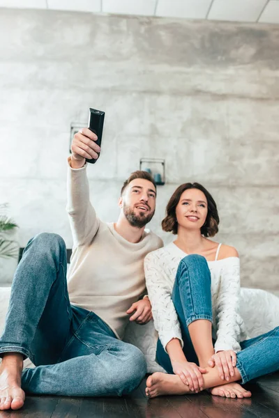 Vista de ángulo bajo del hombre feliz sentado en el suelo cerca de la mujer alegre y la celebración de mando a distancia - foto de stock