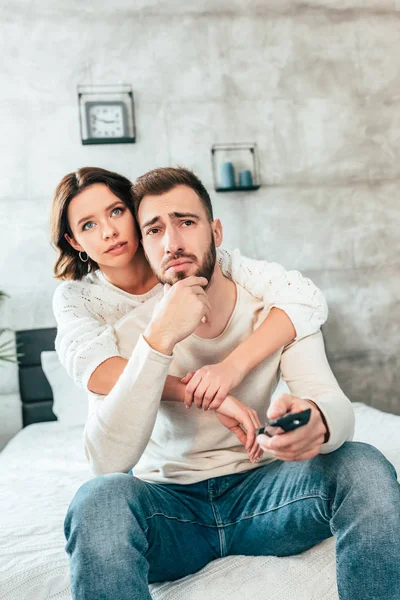 Brünette Frau umarmt aufgebrachten Mann, der auf Bett sitzt und Fernbedienung in der Hand hält — Stockfoto