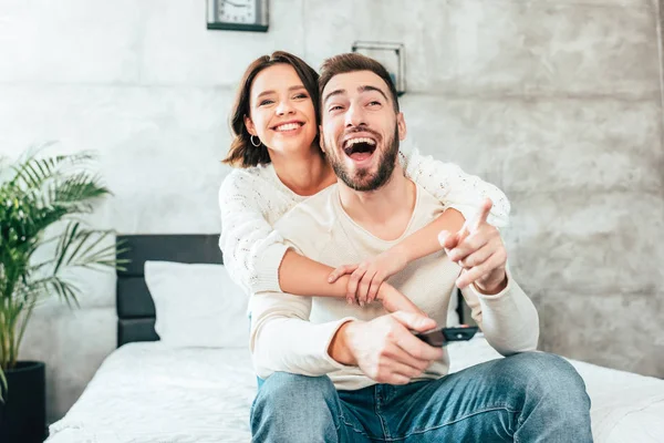 Brunette woman hugging happy man sitting on bed and holding remote controller and pointing with finger while watching movie — Stock Photo