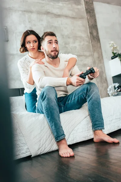 Low angle view of brunette woman hugging handsome man sitting on bed and playing video game — Stock Photo
