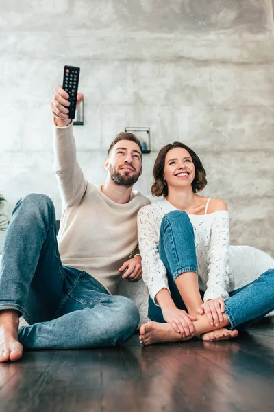 Niedrigwinkel-Ansicht eines gutaussehenden Mannes, der auf dem Boden neben einer glücklichen Frau sitzt und eine Fernbedienung in der Hand hält — Stockfoto