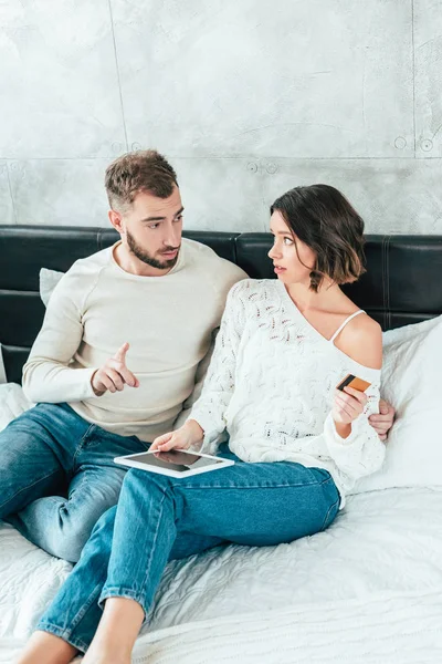 Hombre guapo mirando mujer atractiva sosteniendo tarjeta de crédito y señalando con el dedo cerca de la tableta digital - foto de stock