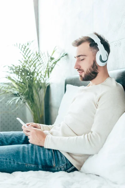 Homem barbudo bonito usando smartphone enquanto ouve música em fones de ouvido — Fotografia de Stock