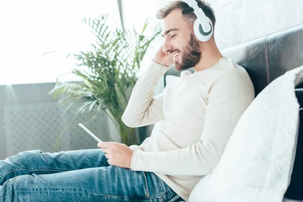 Hombre barbudo feliz usando el teléfono inteligente mientras escucha música en los auriculares - foto de stock