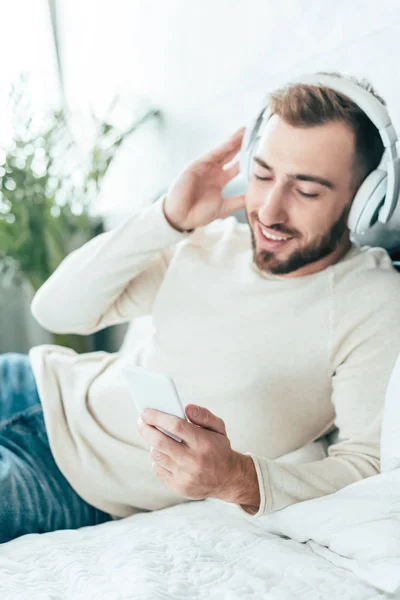 Hombre barbudo alegre usando teléfono inteligente mientras escucha música en los auriculares — Stock Photo