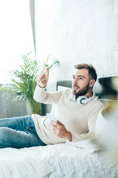 Selective focus of handsome bearded man in headphones holding smartphone and pointing with finger in bedroom — Stock Photo