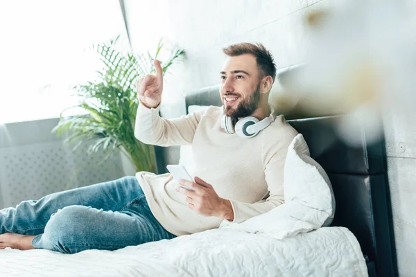 Enfoque selectivo de hombre barbudo feliz en auriculares que sostienen el teléfono inteligente y señalan con el dedo en el dormitorio - foto de stock