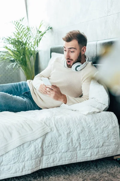 Selective focus of surprised man  in headphones looking at smartphone in bedroom — Stock Photo
